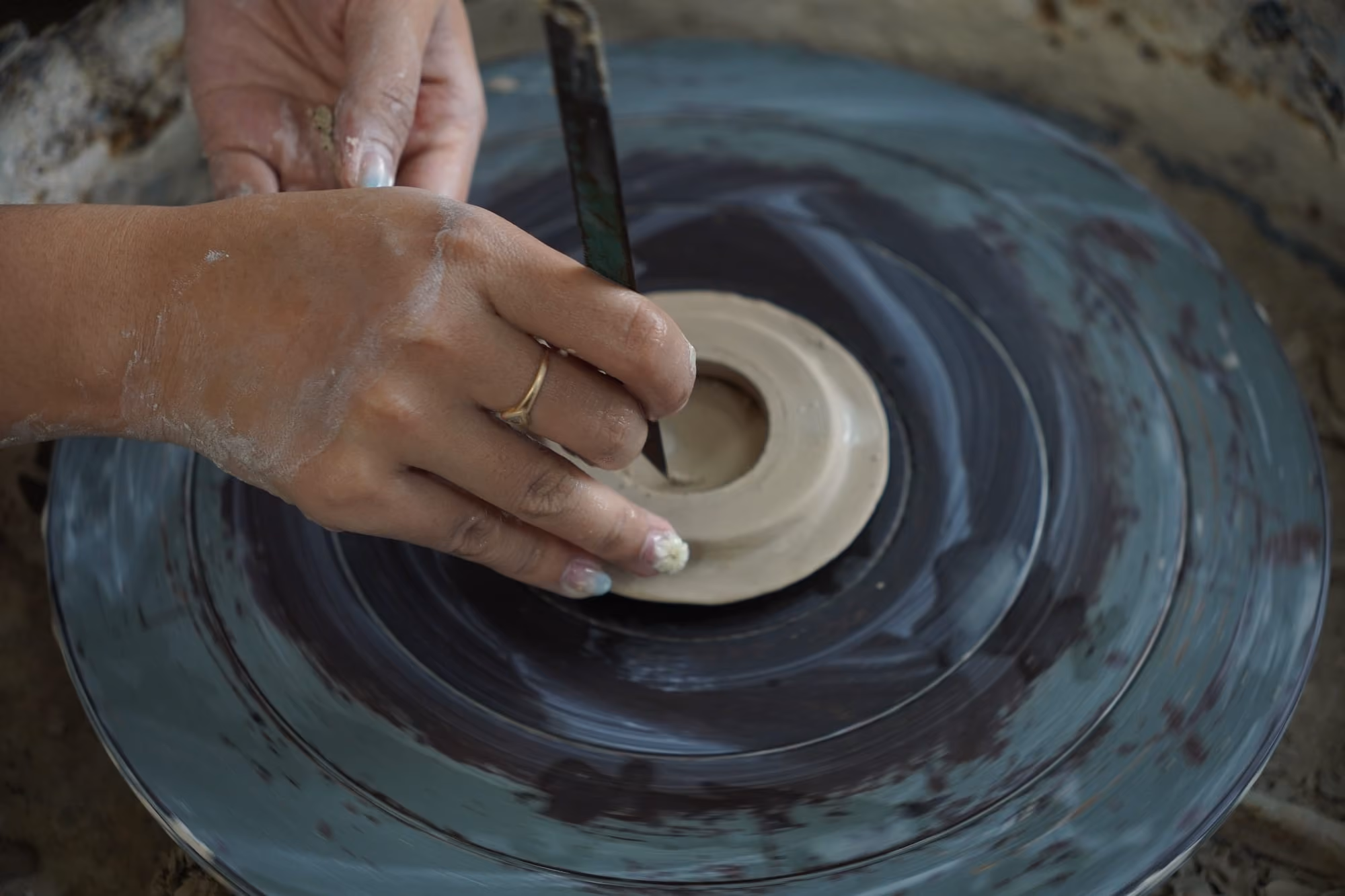A hand with a wooden tool smoothing the inner edge of a clay lid on a potter’s wheel