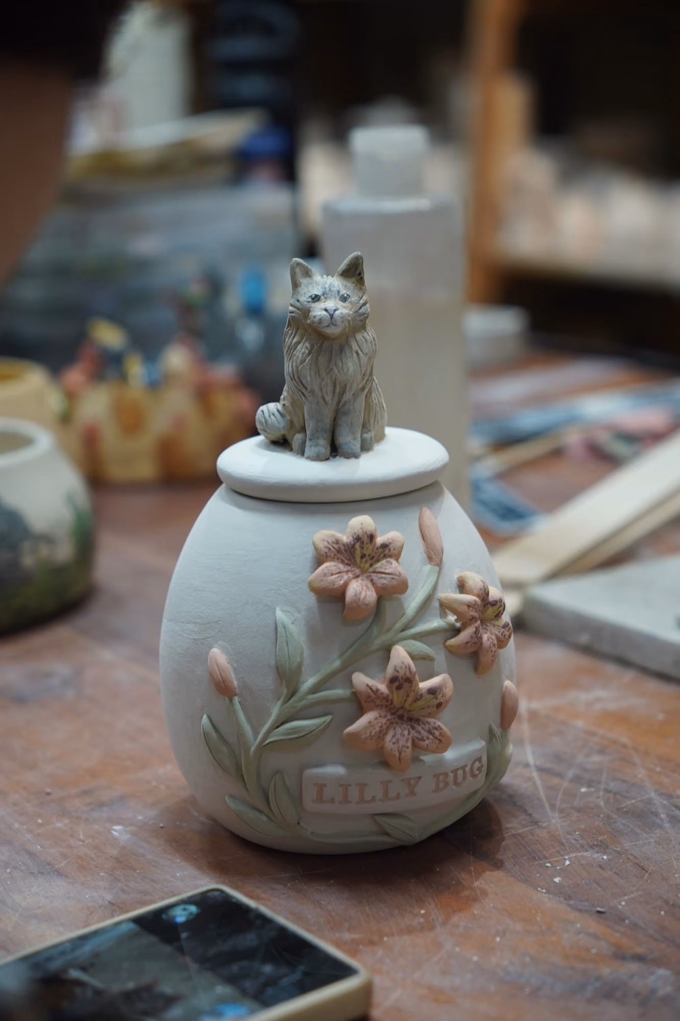 A pale ceramic urn decorated with pink lilies in relief and a small grey cat figurine sitting on the lid, on a cluttered workshop table