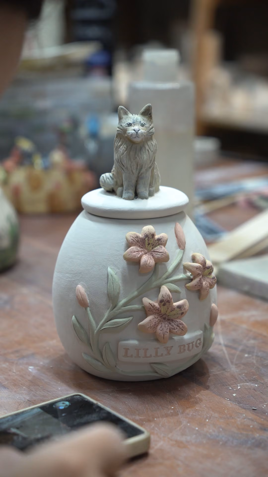 A pale urn with pink relief lilies and a grey cat figurine on its lid, filmed close up on the workshop table