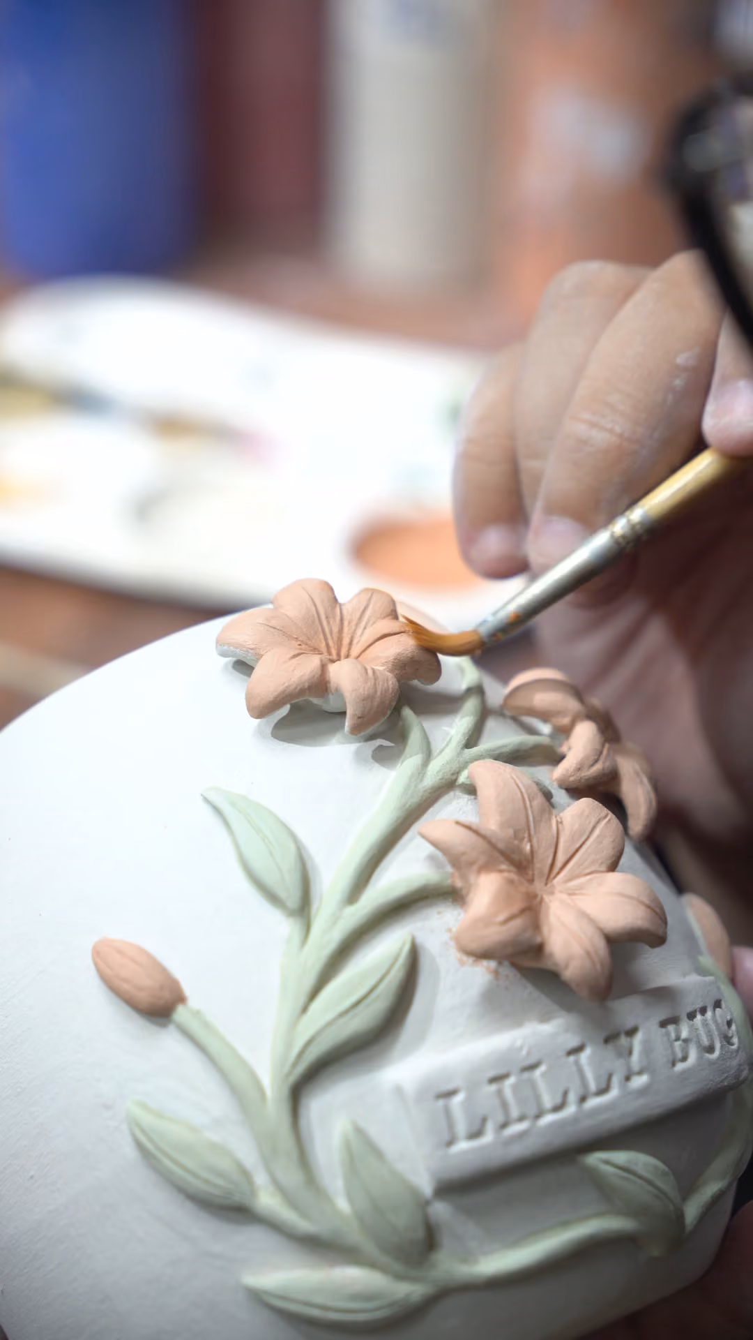 A thin brush painting pink colour onto relief lilies on the side of a white urn