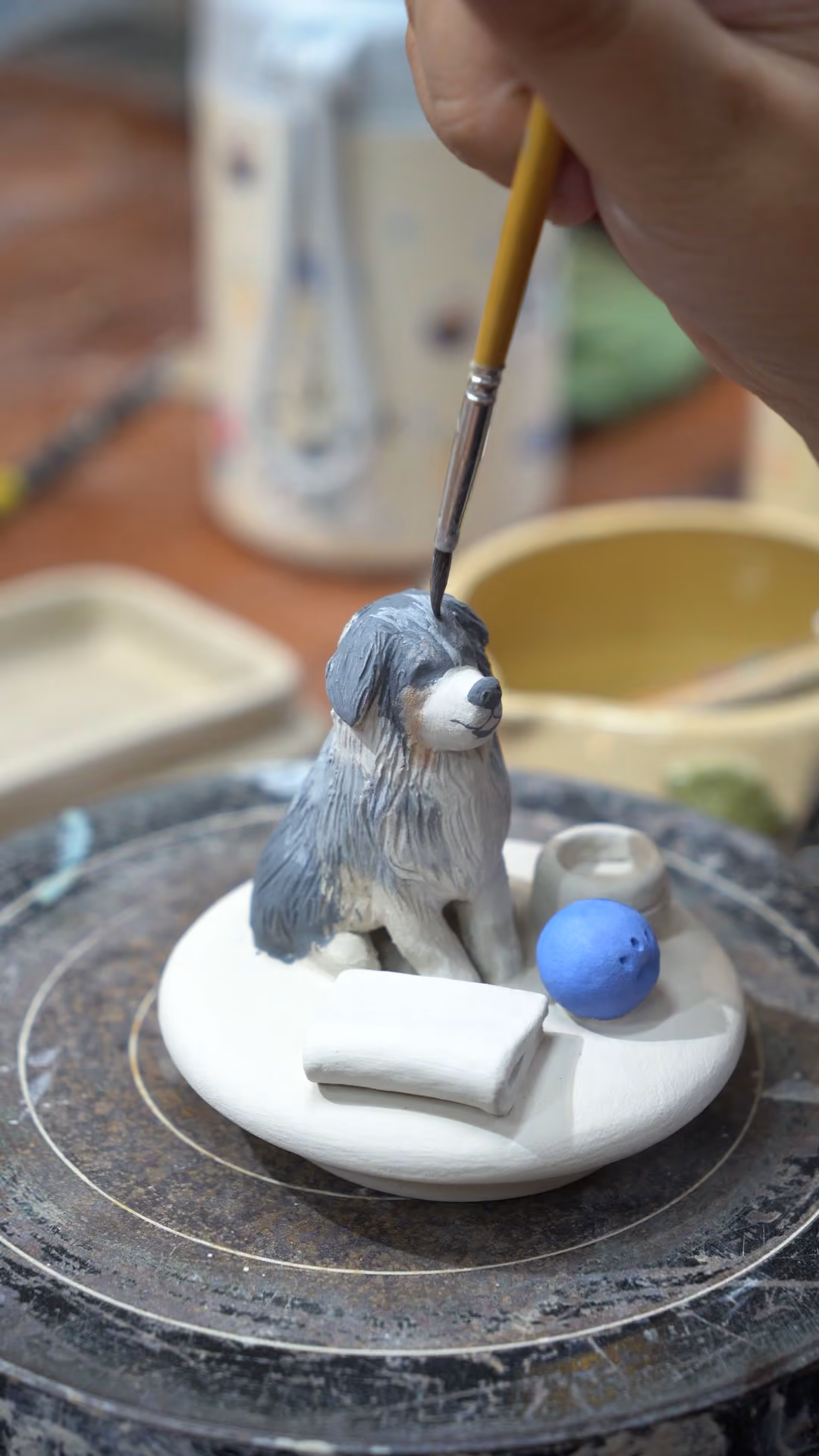 A brush adding grey colour to a long-haired dog figurine sitting on a white lid beside a blue ball