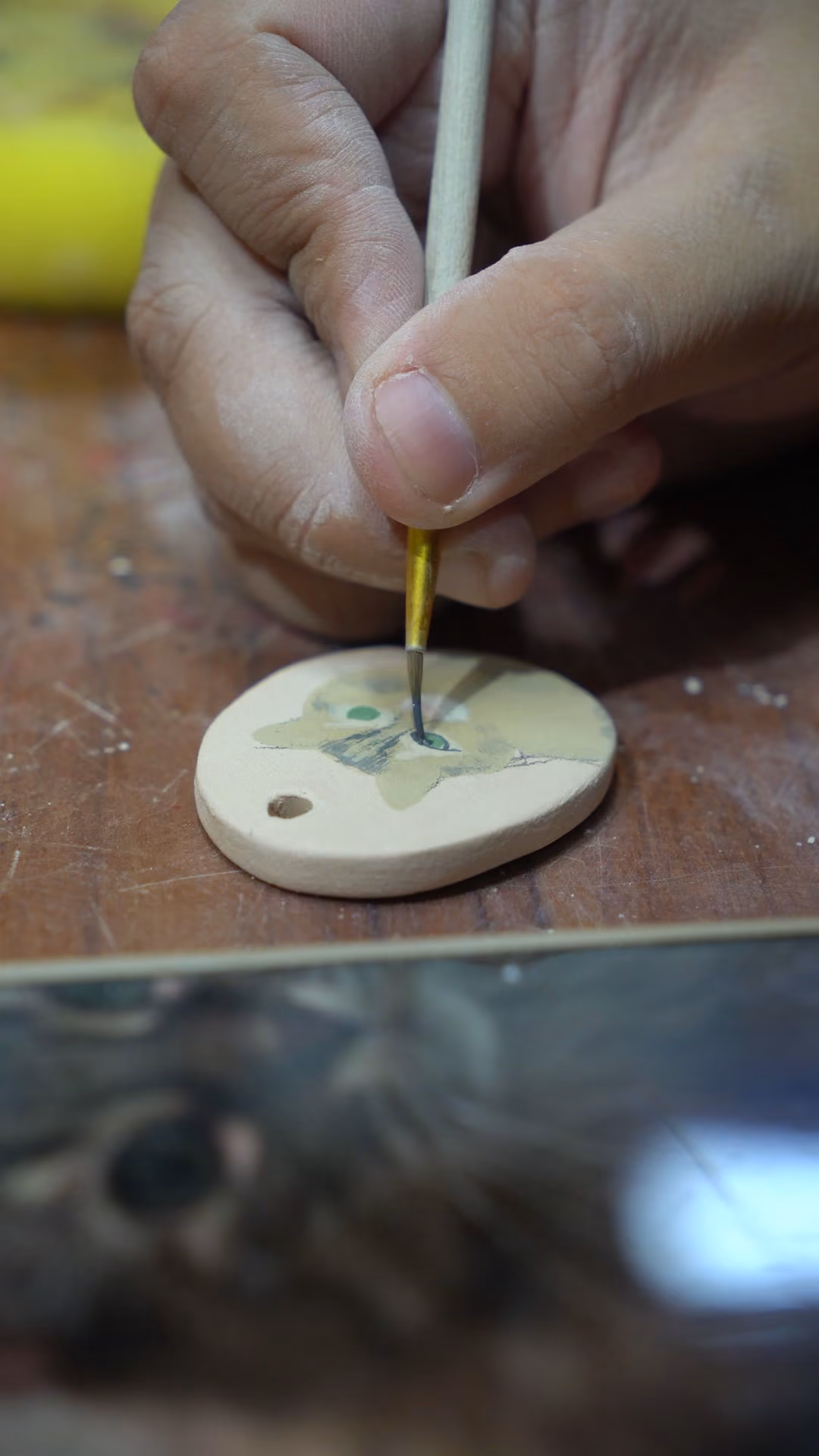 A very thin brush painting detail onto a small round ceramic keepsake held flat on the table