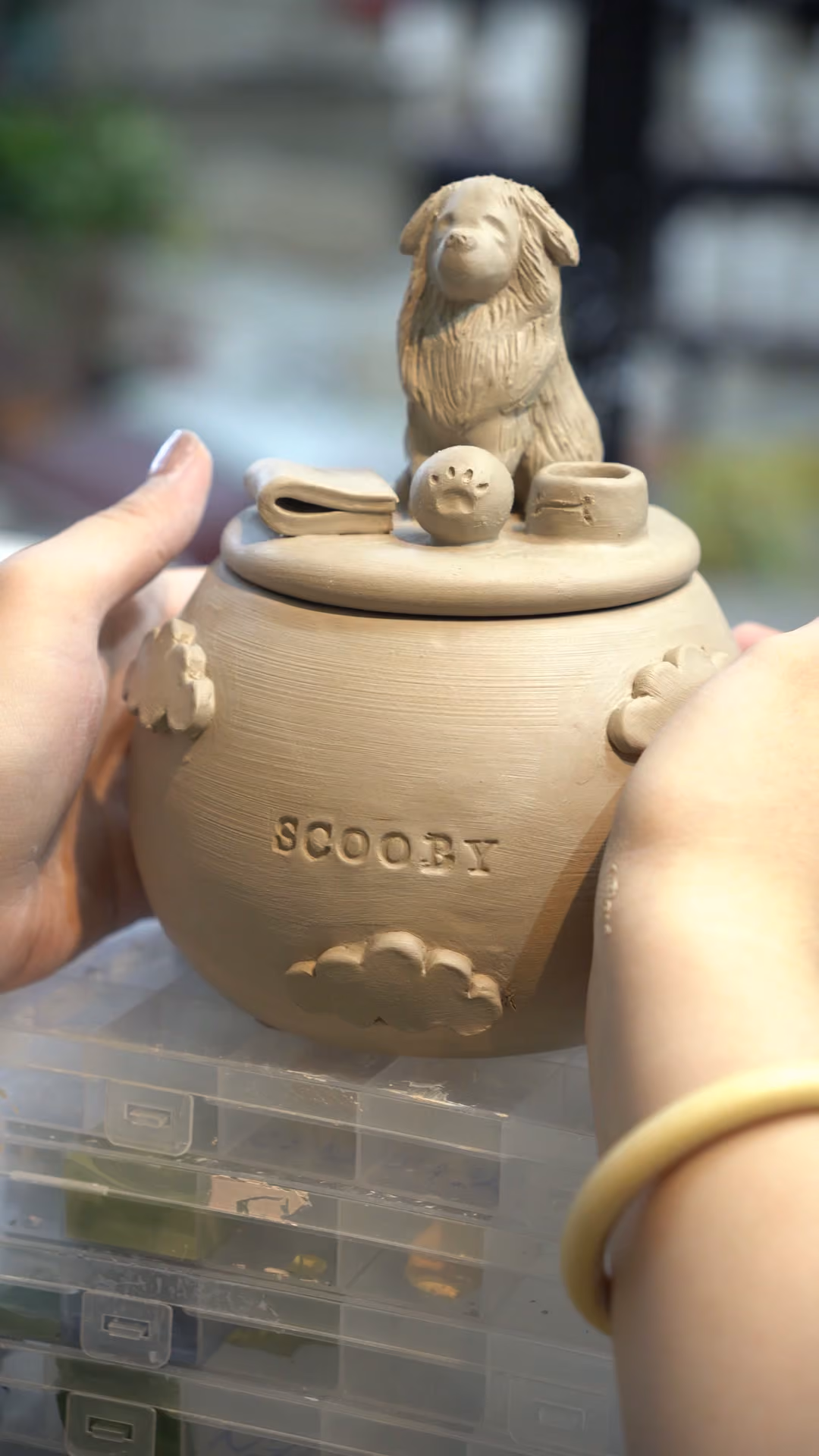An unfired tan clay urn engraved with the name Scooby, with a sculpted dog on the lid, on a workshop table