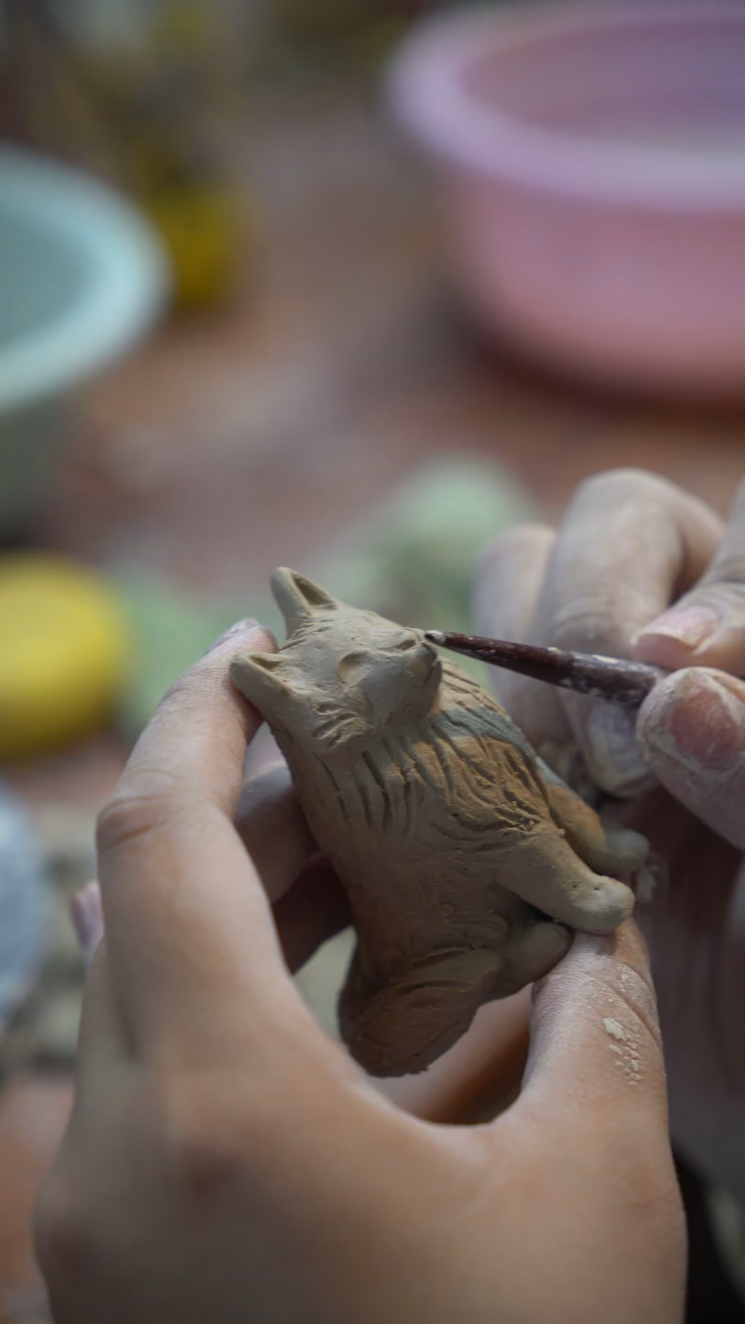 Hands holding a raw clay animal figurine while a pointed tool draws fur texture along its back