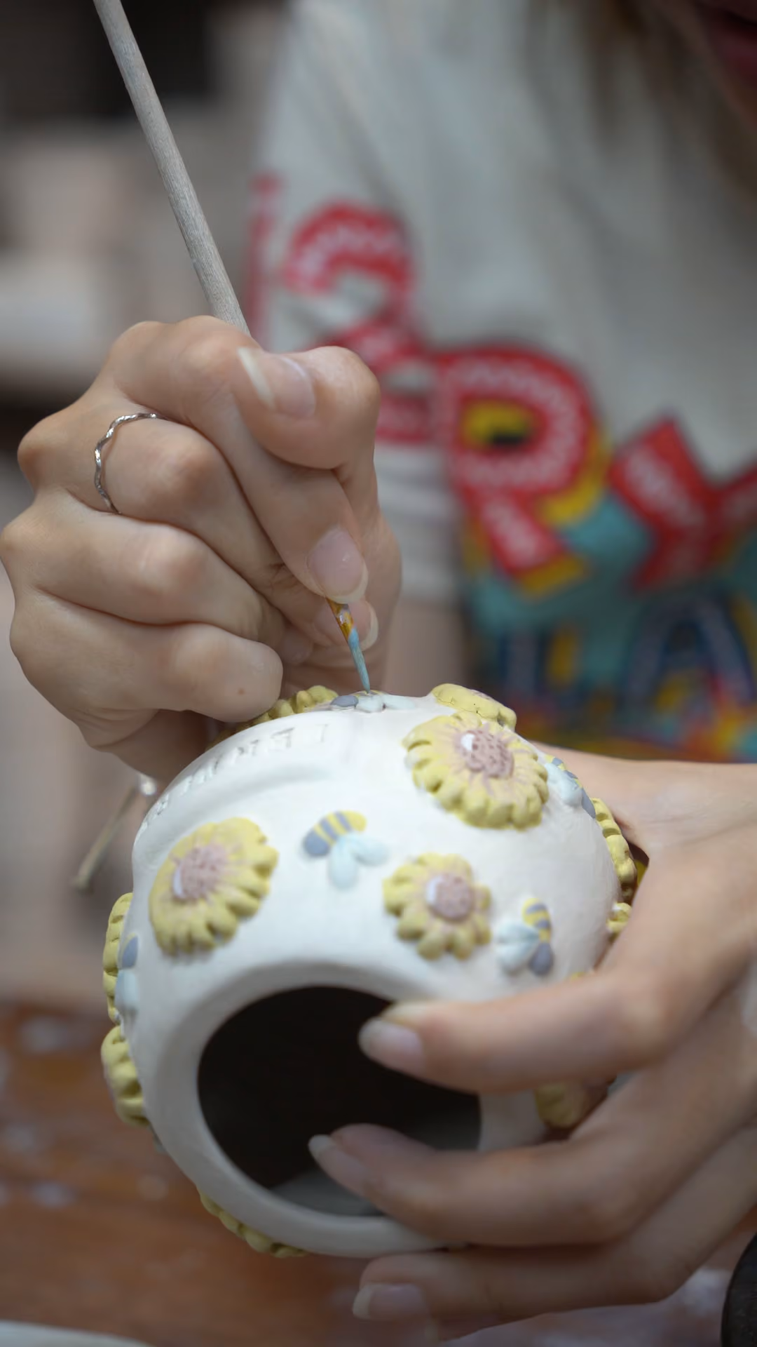 A hand holding a round urn covered in relief sunflowers while another hand paints the petals yellow with a thin brush