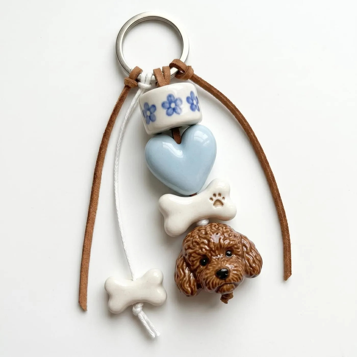 A finished keychain on a split ring: a sculpted poodle head below a ceramic bone, a pale blue heart and a flower-patterned bead, strung on a leather cord