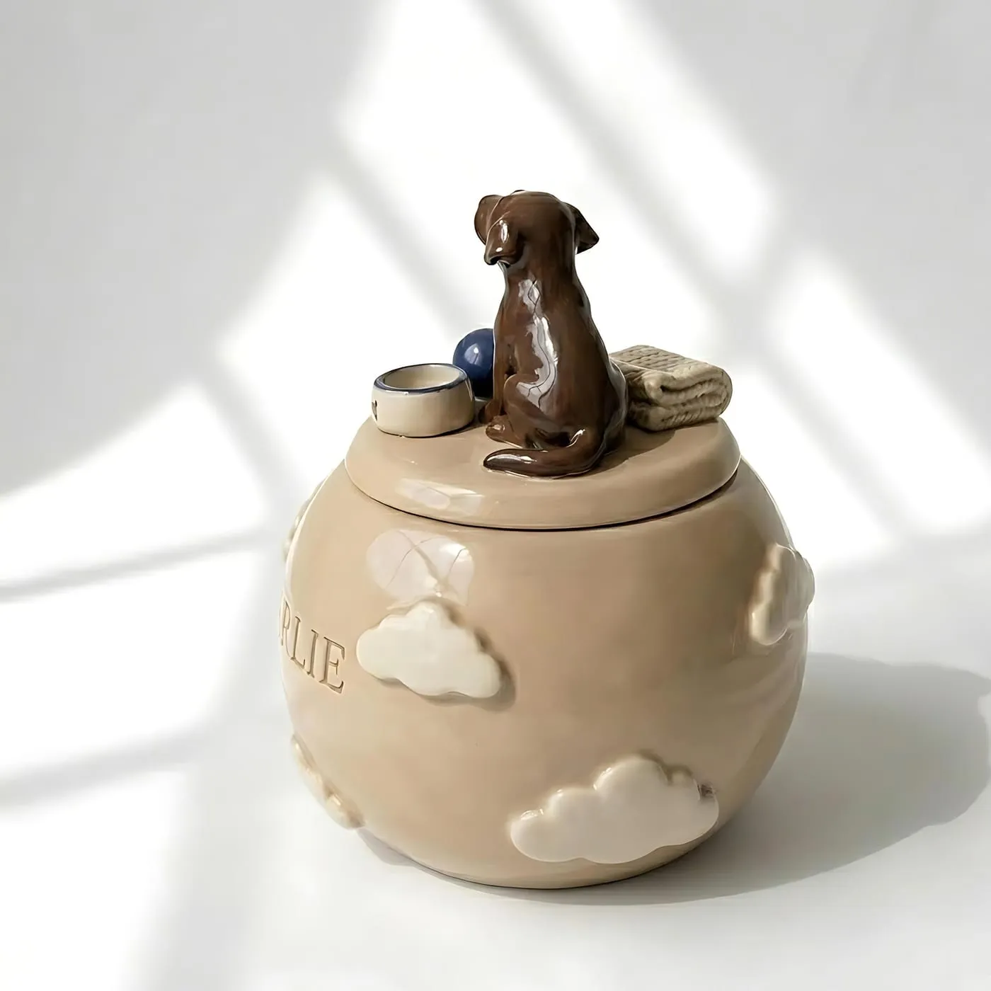 A pale ceramic pet urn with raised cloud motifs and a sculpted brown dog on the lid, with the seam between lid and body clearly visible