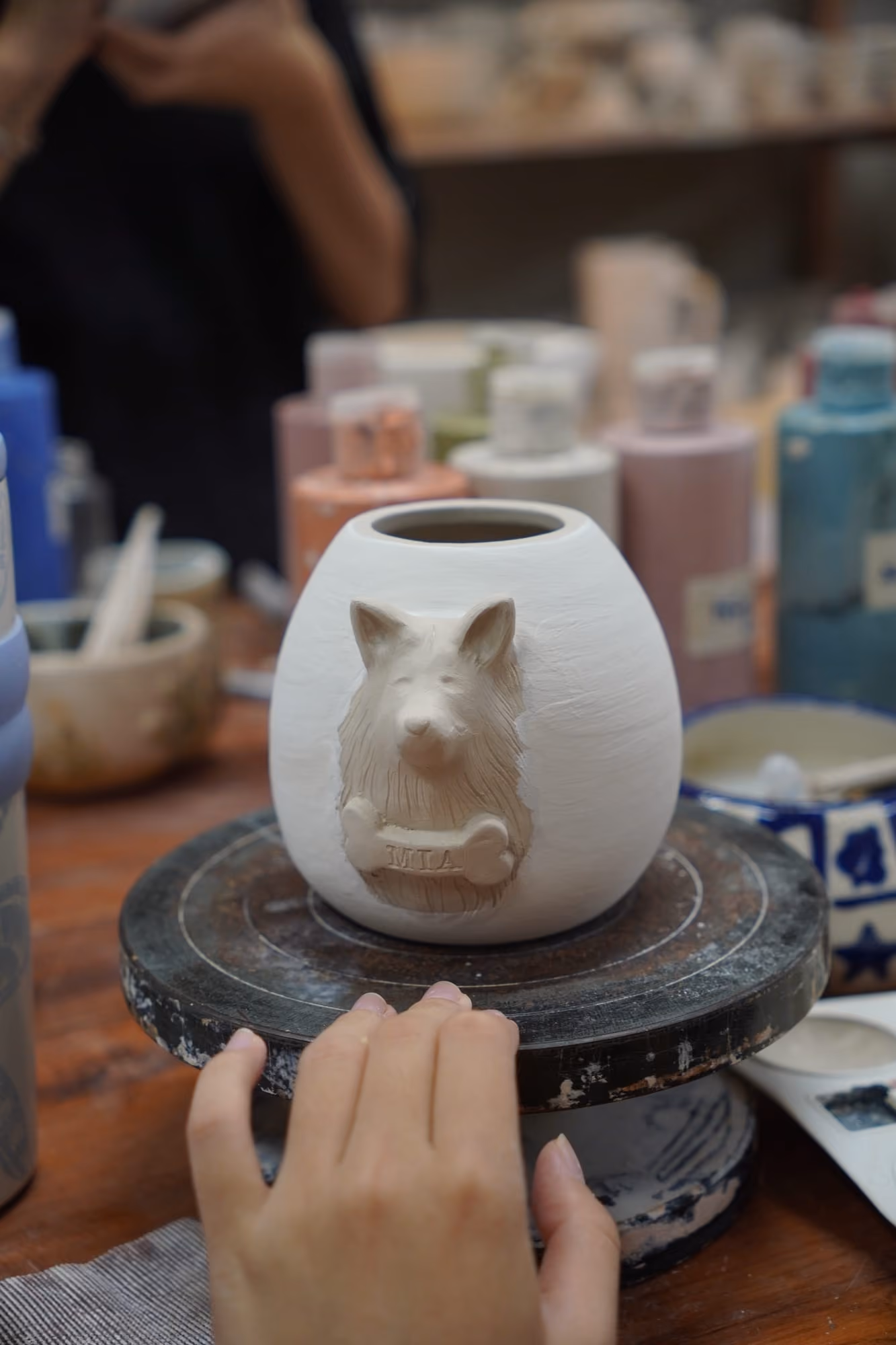A white clay urn on a banding wheel with a pointed-eared animal face sculpted in relief on its side