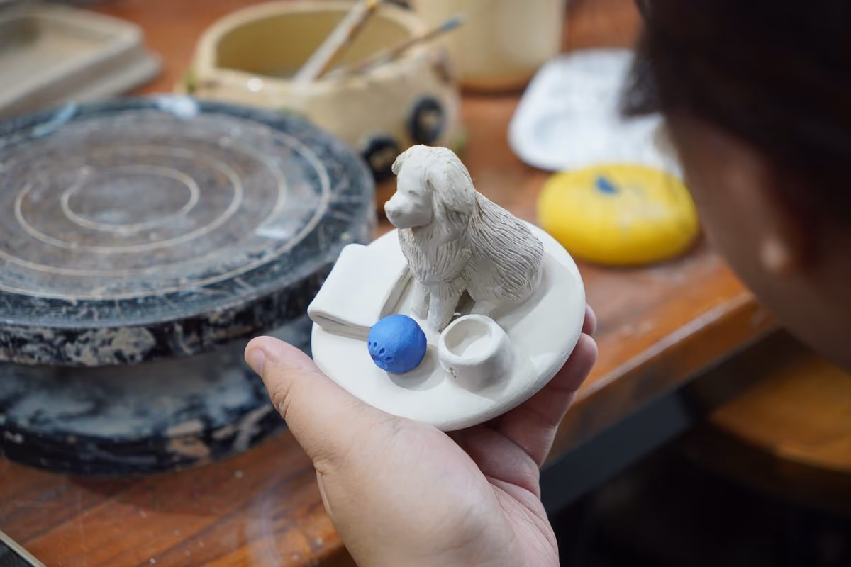 A hand holding an unfired white clay urn lid with a sculpted long-haired dog sitting on it beside a folded blanket, a blue ball and a small bowl
