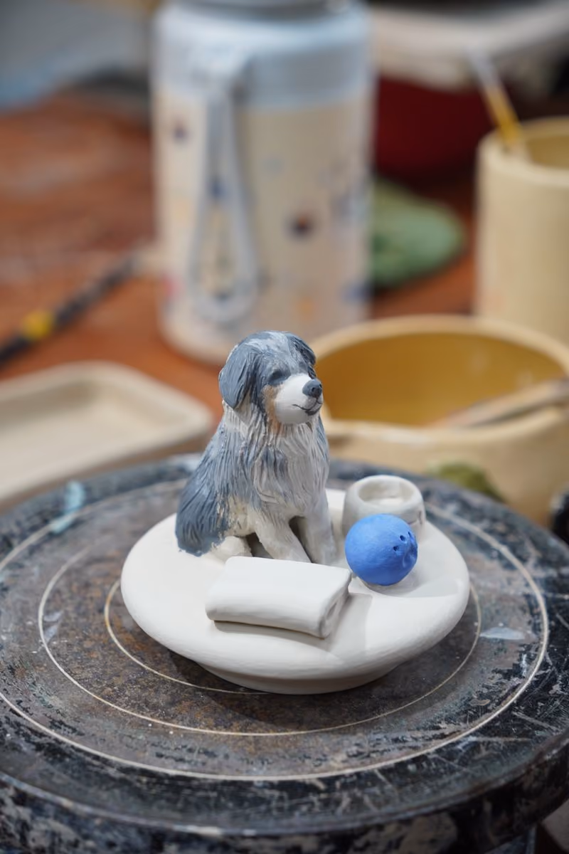 The same lid on a banding wheel after colour has been added, the dog painted grey, white and tan beside the folded blanket, the blue ball and the bowl