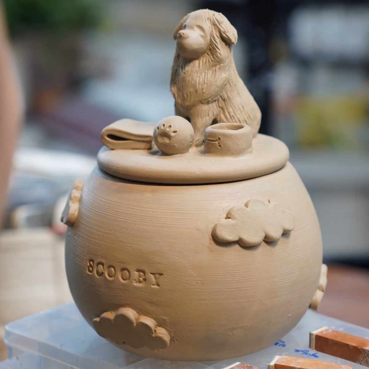 An unfired tan clay urn engraved with the name Scooby, decorated with clouds, with a sculpted dog sitting on the lid