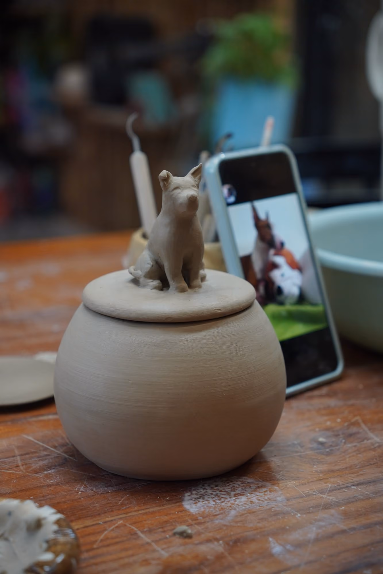 An unfired grey clay urn with a small sitting dog on its lid, next to a phone showing a photo of a dog