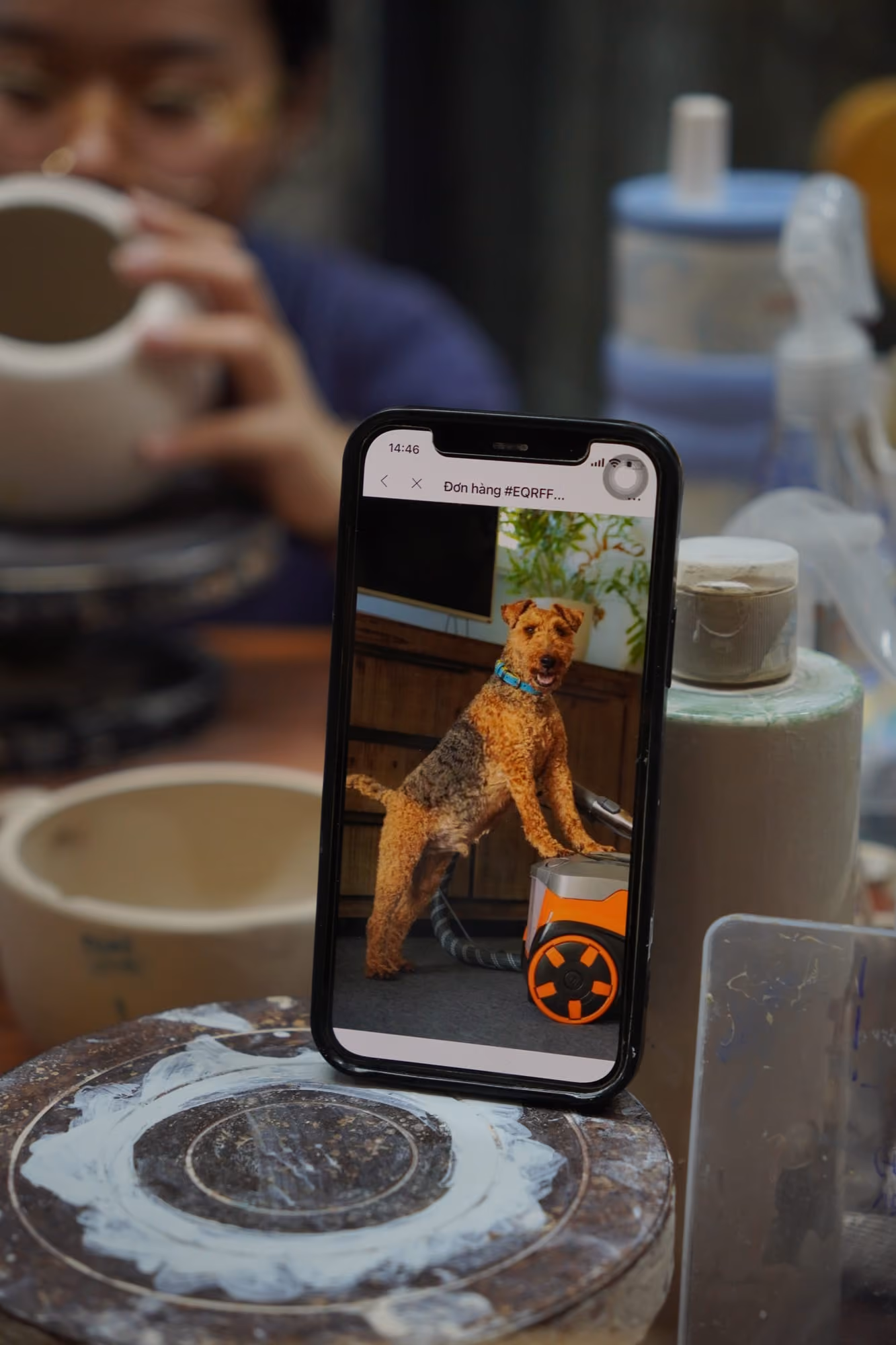 A phone propped upright beside a banding wheel, its screen showing a photo of a brown and black terrier