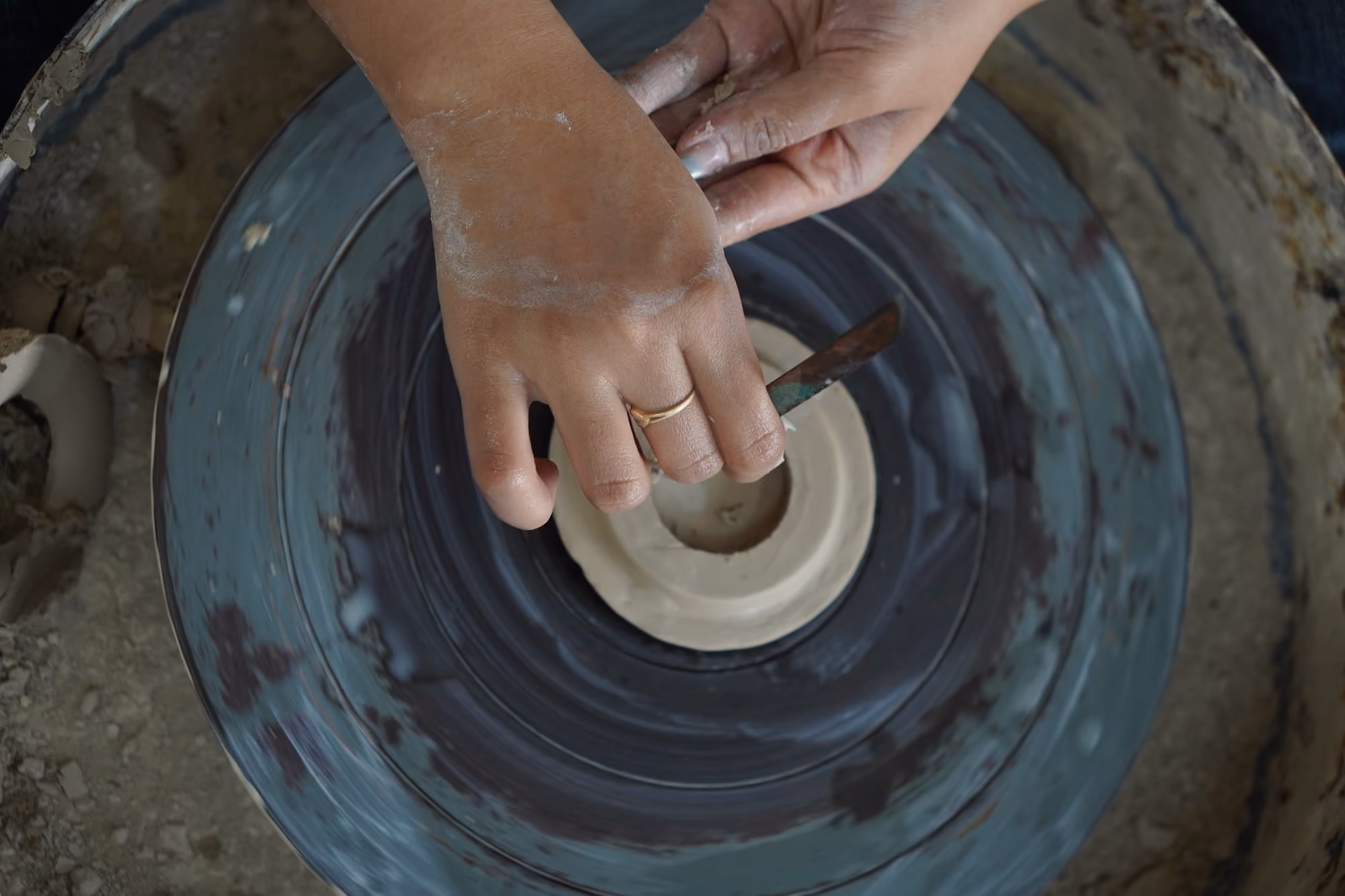 Hands carving a clay disc with a metal tool on a turning potter’s wheel