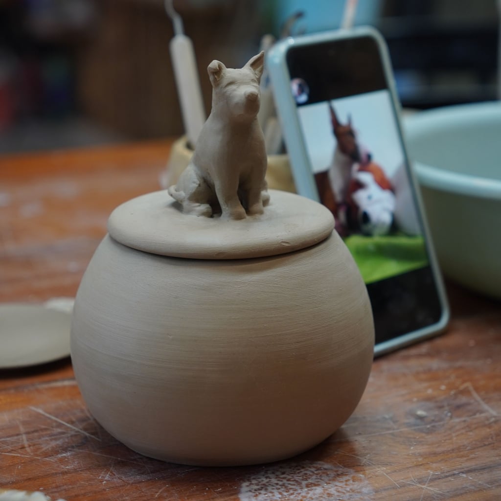 Unfired clay pet urn with sculpted dog figurine photographed next to the customer’s reference photo before firing