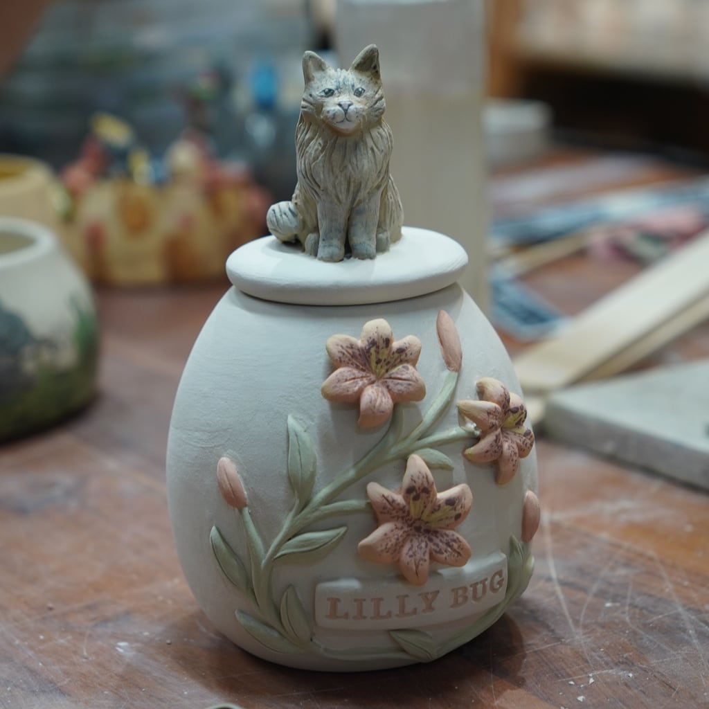 Hand-painted lilies and cat figurine on a ceramic pet urn, glazed and ready for the 1200°C firing